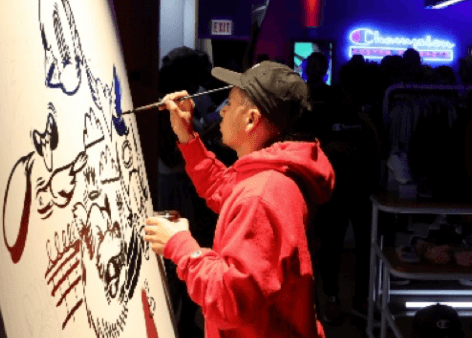 Photograph of a man painting a cartoon image on a large canvas. He is wearing a red sweatshirt and black baseball cap.