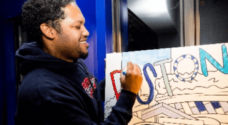 Photograph of a smiling man drawing a cartoon image of the word Boston. He is wearing a sweatshirt.
