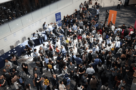 Photographic image that is looking down on a group of around 100 people at a business event. It may be a networking conference. They are all wearing name badges. There are tables with information for people to pick up and read.