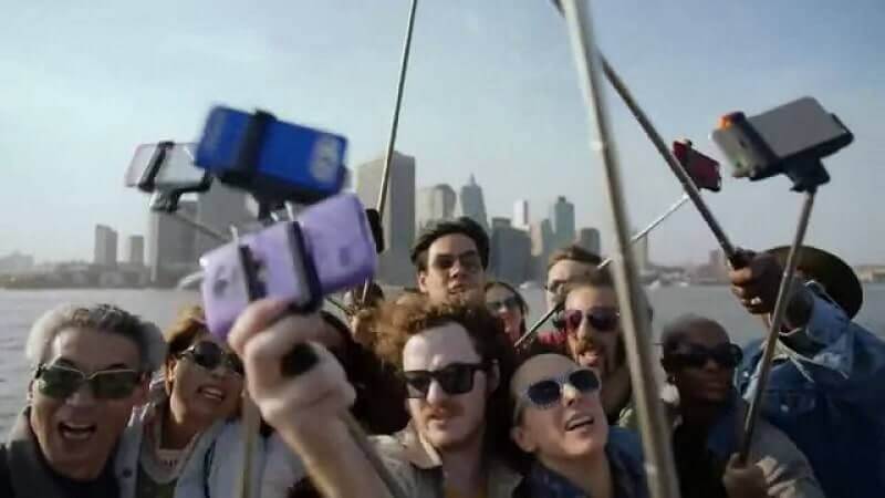 Photograph of a group of around 10 people using selfie sticks. They are taking selfies of themselves in front of the New York skyline. They are all wearing sunglasses.