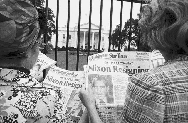 Two women in front of the White House reading about the Watergate scandal. It is the 1970s. The photograph is black and white.