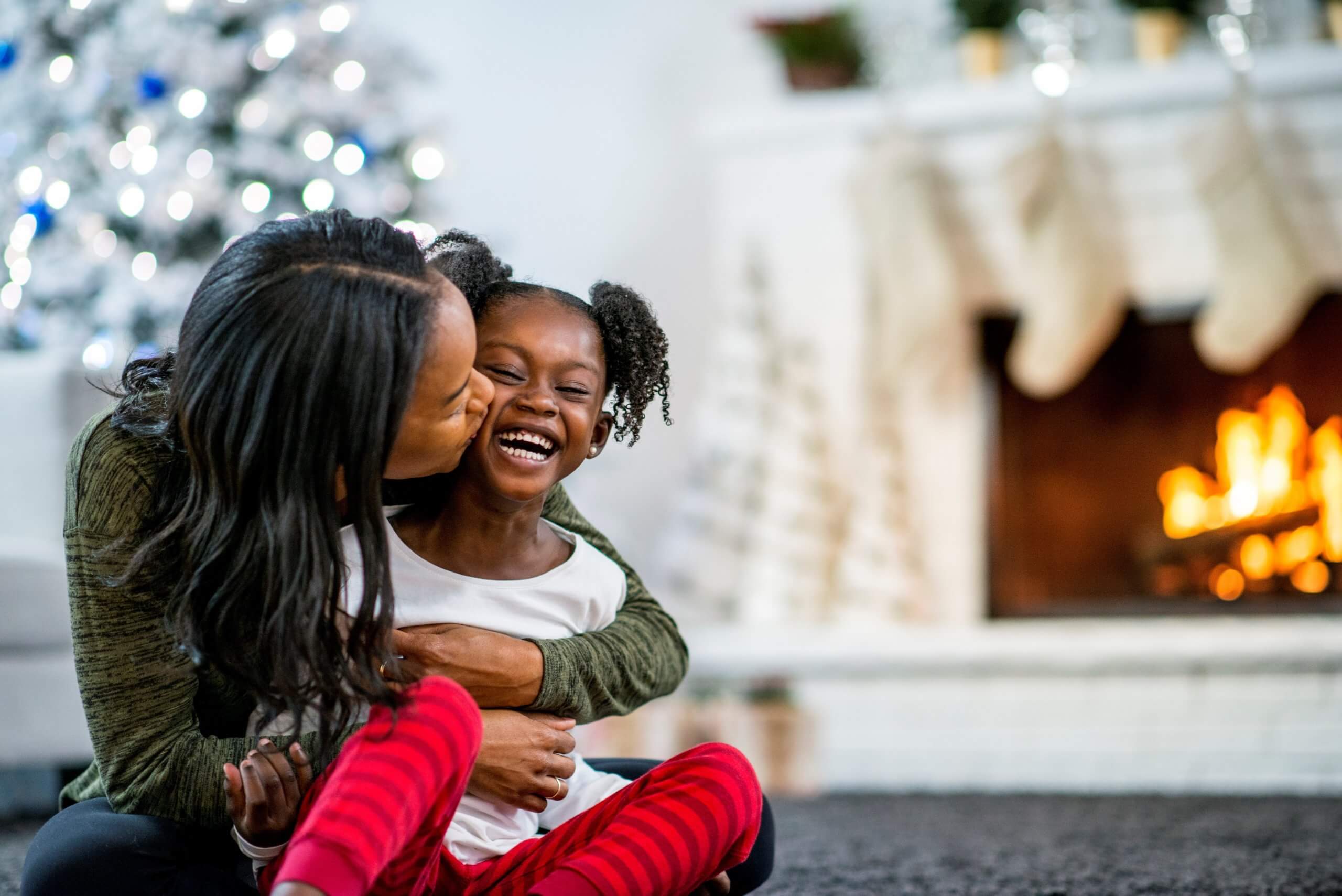 Photograph of mother and daugher hugging in front of a Christmas tree. There is a log fire with stocking hanging up in the background of the picture.