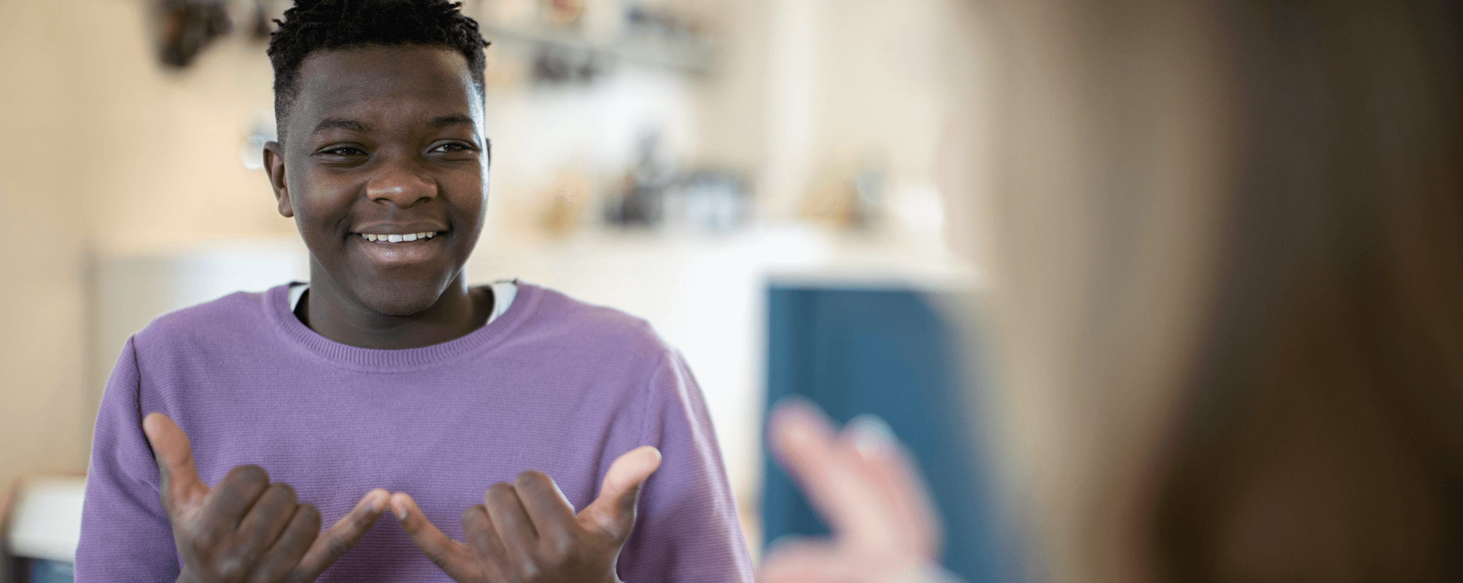 Jeune homme noir communiquant en utilisant le langage des signes. Il sourit. Il se trouve à gauche de l'écran et porte un haut violet.