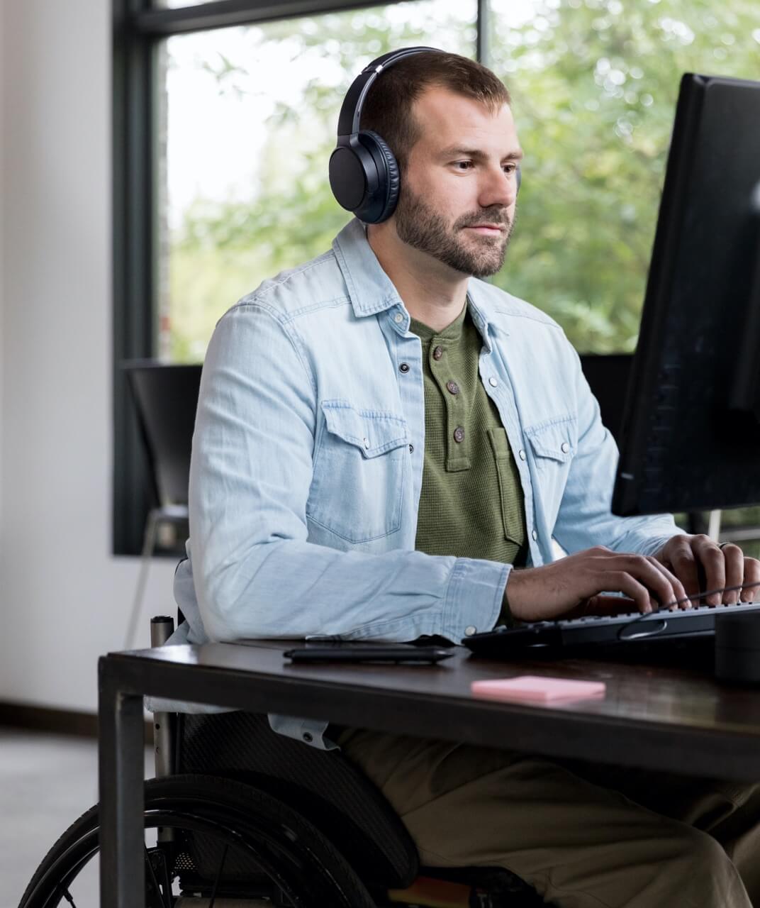 坐在轮椅上的男子坐在电脑前工作。他留着胡须，戴着耳机。他正在键盘上打字，似乎是在办公室工作。