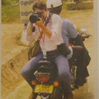 Man on the back of a motorbike pointing a long range camera lens at the photographer.