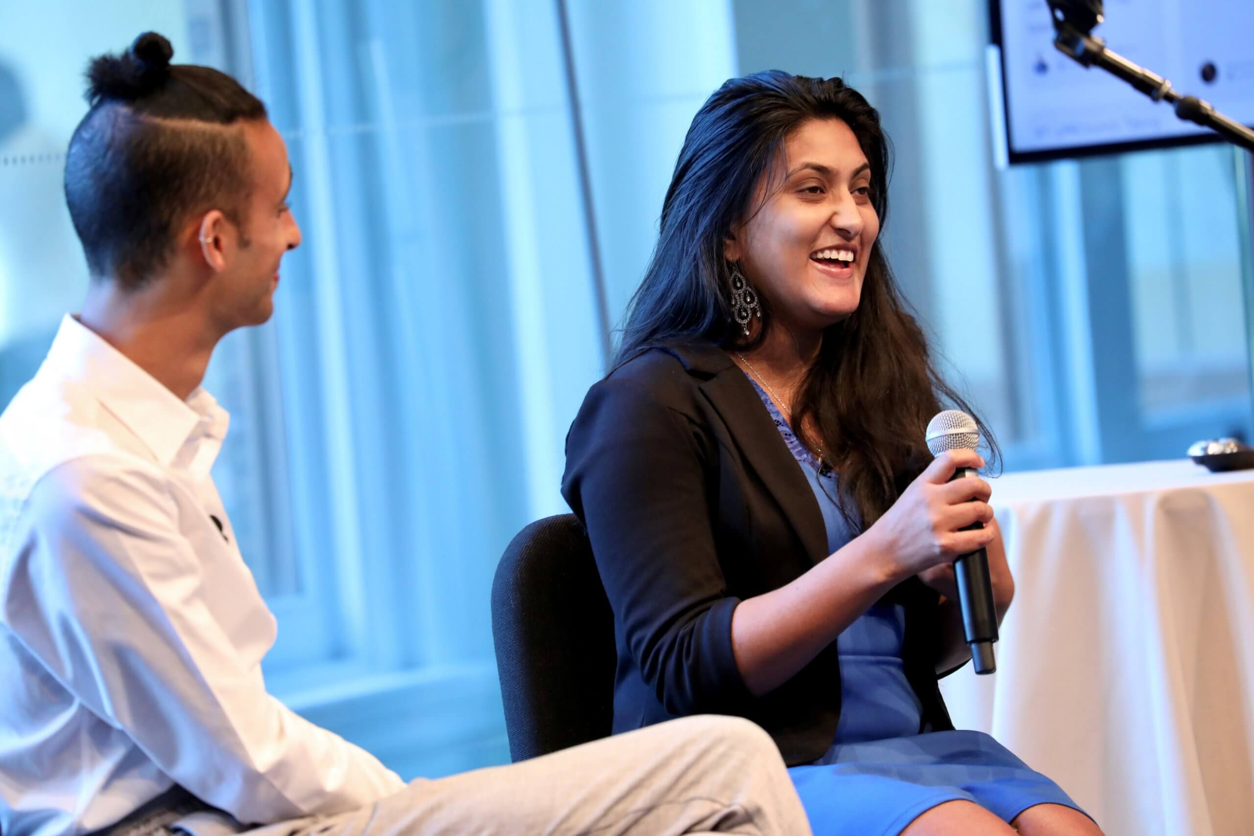 A man and woman talking at a conference. Sena Pottackal is the woman talking on the right hand side. She is holding a microphone.