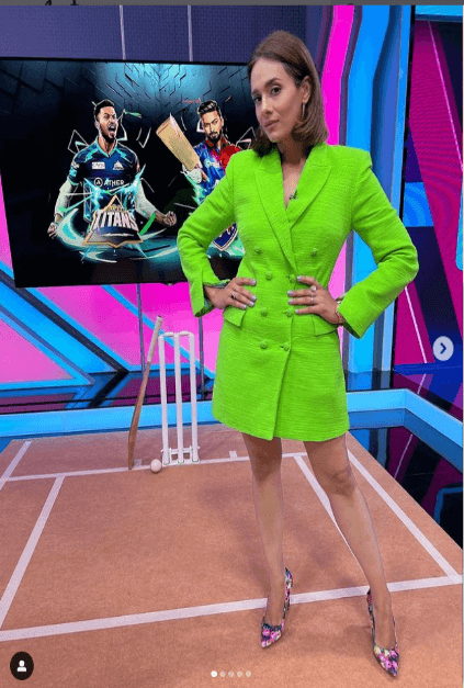 Mayanti Langer, a sports broadcaster from India is wearing a green dress and is standing before a giant TV screen showing an image of two men.