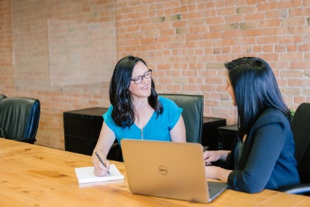 Eine Frau in einem blaugrünen Hemd sitzt neben einer Frau in einem dunklen Blazer und lächelt sich gegenseitig an, während sie über ihre Arbeit sprechen.
