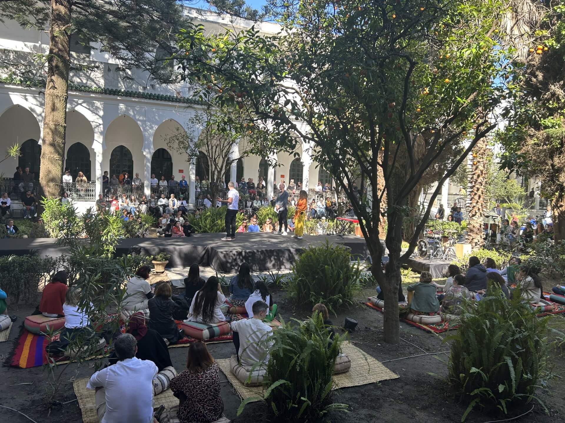 A group of some 30 plus people sat on Morroccan-style cushions in an open air courtyard in Tangiers. They are listening to speakers on a central stage. There are beautiful trees growing in the courtyard.