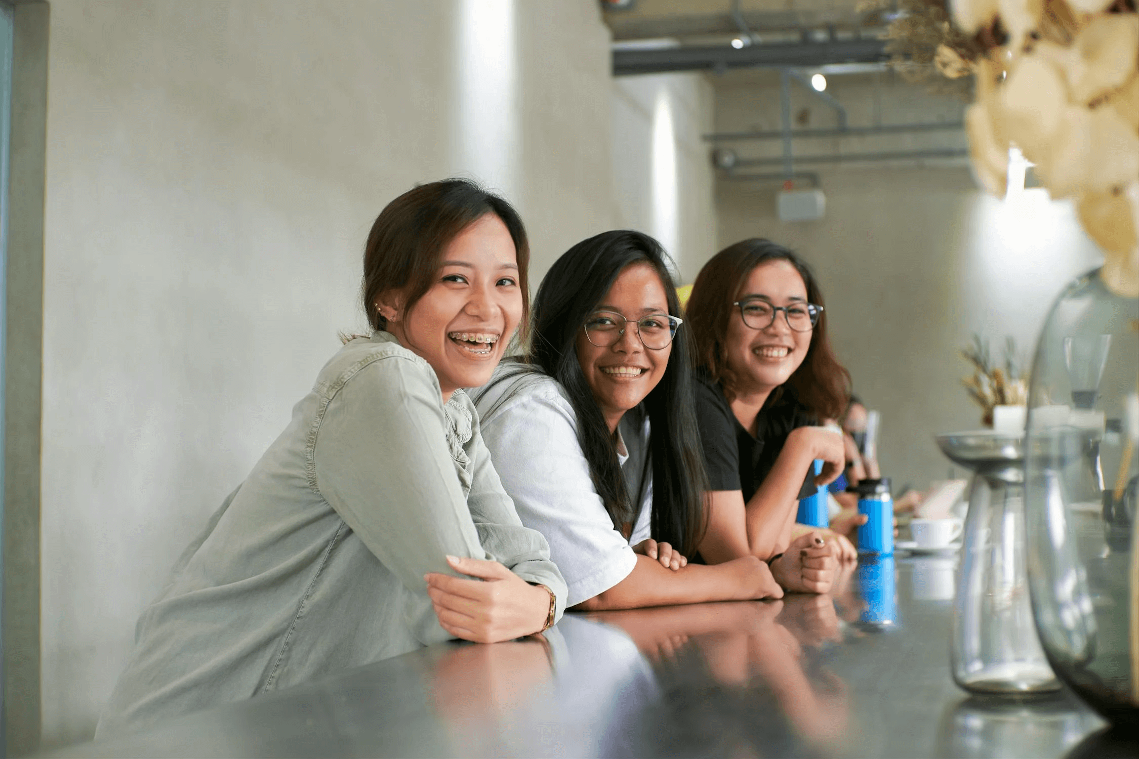 Um terreno diversificado de três mulheres asiáticas, sentadas a uma mesa e a sorrir para a câmara.