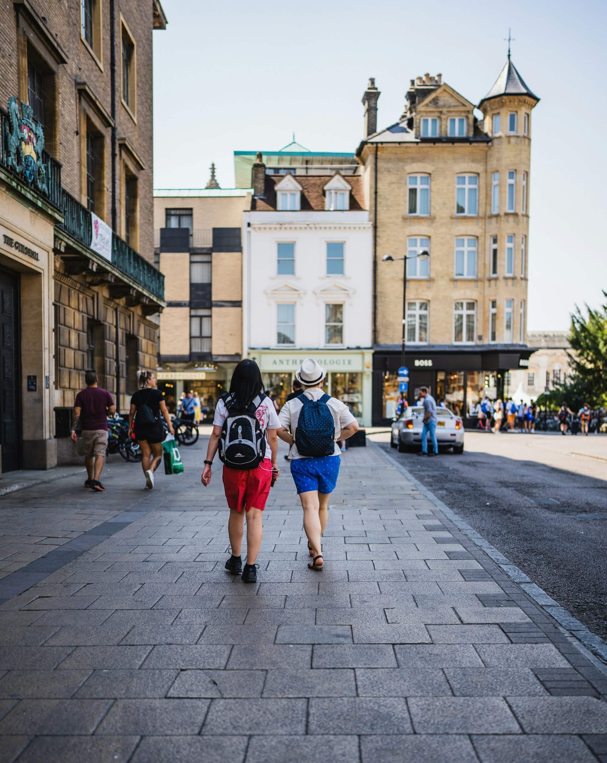 The rear view of a young couple walking through the streets of a European town. The woman on the left wears red shorts, the man on the right wears a hat and small backpack.