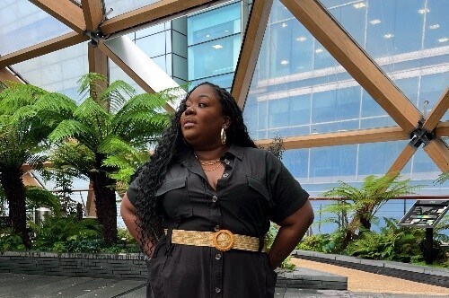 A woman stands confidently outdoors in a modern space with lush greenery and large wooden beams overhead. She wears a black button-down dress with a beige belt and black boots, accessorized with gold hoop earrings and layered necklaces. Her long, wavy hair falls over her shoulders as she gazes to the side with a poised expression. The setting features a sleek, wet stone floor and tall ferns, creating a serene, contemporary atmosphere.