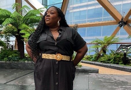 A woman stands confidently outdoors in a modern space with lush greenery and large wooden beams overhead. She wears a black button-down dress with a beige belt and black boots, accessorized with gold hoop earrings and layered necklaces. Her long, wavy hair falls over her shoulders as she gazes to the side with a poised expression. The setting features a sleek, wet stone floor and tall ferns, creating a serene, contemporary atmosphere.