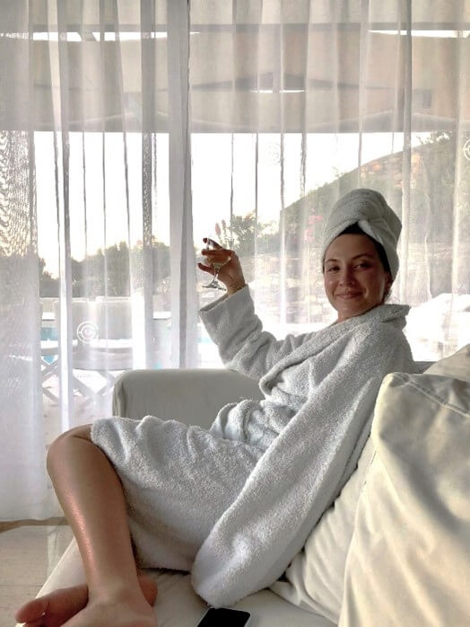 A young woman sitting on the sofa in a holiday villa. She is wearing a white bath robe and hair wrapped in a towel. She is drinking a glass of wine.